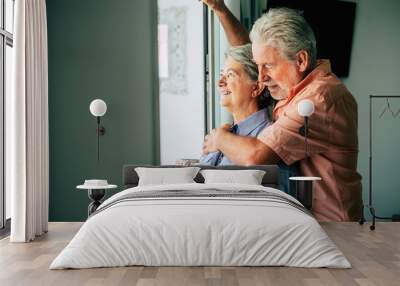 Happy old people caucasian senior couple enjoying home together drinking healthy beverage like tea and looking outside the window together with hug. and love - forever concept Wall mural