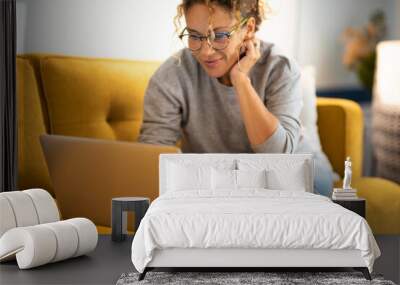 Happy young woman working on laptop sitting on sofa in living room at home. Businesswoman in casuals working from home. Caucasian woman in eyeglasses smiling while using laptop. Wall mural