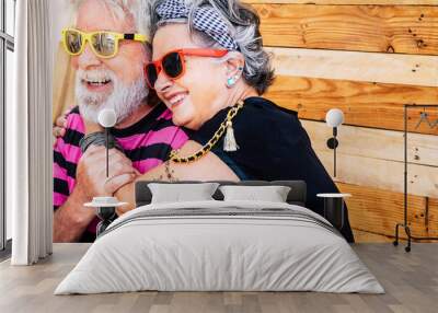 No limit age with nice and funny elderly caucasian couple have fun and smile in a portrait with wooden background - trendy and coloured accessories on old cheerful and happy people Wall mural