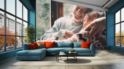 Old senior couple in love hug and embrace with romance together at home with outside view in windows background - happy mature retired people lifestyle enjoying caring each other and smiling Wall mural