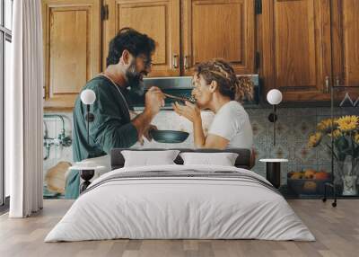 One happy real couple at home enjoying life together and relationship. Man giving food to woman to taste. Male people cooking for his wife. Male and female enjoying leisure activity indoor in kitchen Wall mural