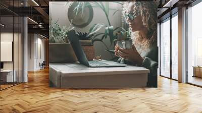 One lady using laptop at home drinking tea or coffee in indoor leisure activity online. Pretty woman working on computer and smiling. Lady surfing the web alone with nature plants garden in background Wall mural