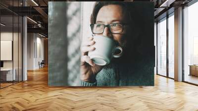 One man with thoughtful and serene expression on face drinking coffee or tea alone at home looking outside the window. Mature male people alone. Healthy mindful lifestyle concept. Wearing glasses Wall mural