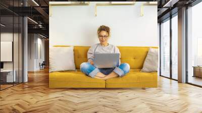 One woman smiling and using laptop computer at home sitting comfortably on a yellow sofa in living room. Smart working female people notebook. Surfing the web. Enjoying technology and connection Wall mural