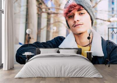 One young guy sitting outdoor with city buildings in background looking on camera with trendy hair and clothes. Leisure activity. Modern teen male posing outside and smiling confident Wall mural