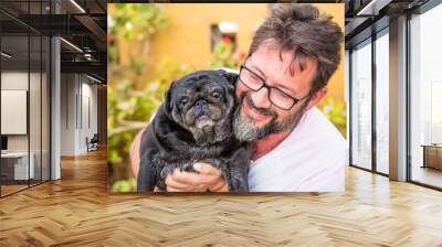 Outdoor nice portrait of adult caucasian man with black beard and same color old funny dog pug - garden in background and best friend love concept Wall mural