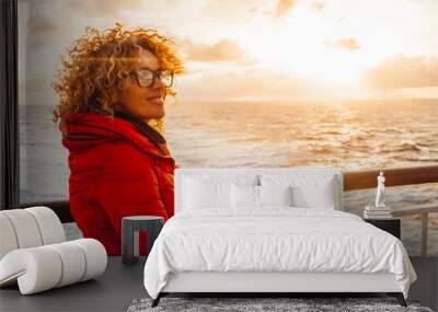 Portrait of a cheerful young adult woman enjoying sunset outside traveling the ocean on a ferry cruise ship boat alone. Traveler people lifestyle, Red jacket. Orange warm sky background. Transport Wall mural