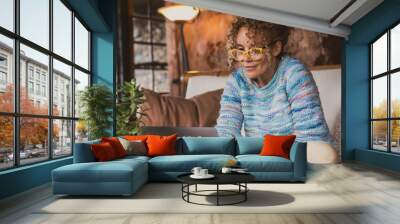 Portrait of happy modern woman at home sitting on the sofa using computer and wearing eyewear. Female people surf the web and work with laptop smiling. Indoor technology leisure activity concept Wall mural