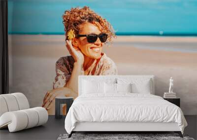 Portrait of happy tourist sitting and smiling at the beach with blue sky and ocean in background. Travel and tourism in summer holiday vacation. Female people with sunglasses on the sand Wall mural