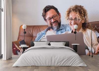 Portrait of two people man and woman enjoy technolofy connection toether with laptop and phone smiling and watching the computer - happy cheerful couple at home sitting on the sofa at work Wall mural