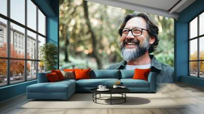 Portrait of young mature man alone smiling outside with nature trees in background. Environment. Happy people enjoying outdoor leisure activity. Nature feeling. Bearded adult male wearing eyeglasses Wall mural