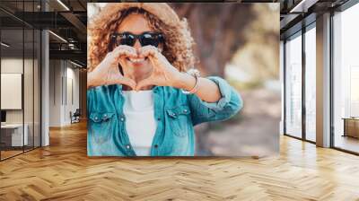 Pretty trendy young woman smile and have fun doing heart gesture sign symbol with hands to the camera. Outdoor leisure activity and trees in background. Happy people in tourism and travel lifestyle Wall mural