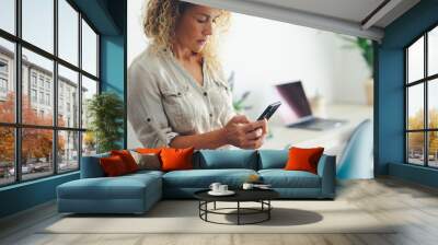 Pretty young adult woman sending message on mobile phone. Concept of social connection with smartphone. Female people using technology at office. Lady work at home. Modern workplace in background. Wall mural