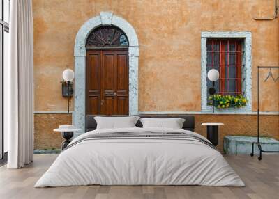 Exterior of an old building with an arched door and a window with a flowering potted plant on an orange wall, Italy Wall mural