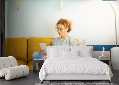 Smart casual woman middle age relaxing at home sitting on yellow sofa and using ereader to read an ebook enjoying indoor leisure activity alone. Sunday time lifestyle. People and technology online Wall mural