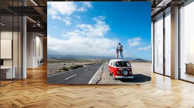 Standing couple on red roof van vehicle hug and love in travel lifestyle - concept of alternative free outdor holiday vacation with young people and blue sky in background Wall mural