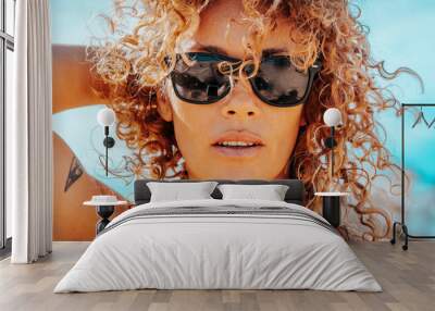 Summer portrait of beautiful young adult woman with sunglasses and healthy blonde long curly hair looking at camera. Attractive brunette with blue sky in background. One cute female people lifestyle Wall mural