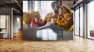 Tenderness time moment for mature caucasian couple at home sitting on the couch together - love and relationship adult people indoor lifestyle - man kissing woman with phone on hand Wall mural