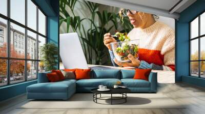 Woman eating a salads with disgusted expression. People on diet and healthy lifestyle habits. Healthy food nutrition concept. Lady tired to eat vegetables. Modern female using computer and eat lunch Wall mural