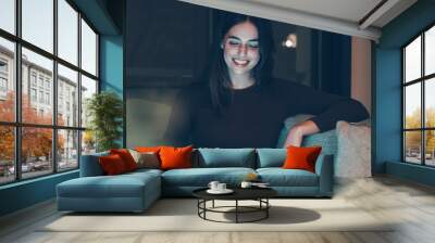 Woman sitting on sofa at night relaxes watching funny videos on her laptop. Pretty female with long hair smiles while she watches funny movie. Wall mural