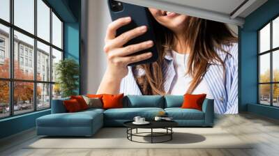 Young, modern woman in casual clothes smiles while using her smartphone at home, scrolling social media or reading digital content, engaged with app downloads and communication Wall mural