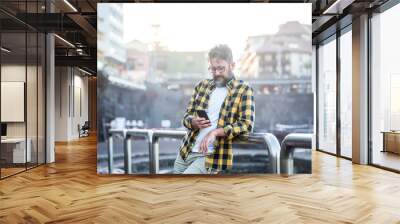 Young adult mature caucasian man using cell mobile phone outdoor with wireless internet free connection. Male people use cellular device in sunny day in the town. City buildings in background Wall mural