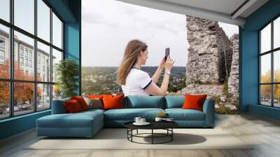 A young woman takes a photo on her phone of a beautiful view of the city from the top of the mountain. Photos and videos on a smartphone. Travel blogger and influencer Wall mural