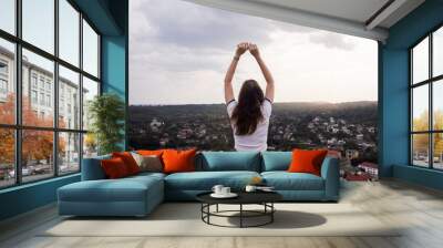 Young woman sits on a mountain with her hands raised like a winner. A gesture of freedom and satisfaction from the seen landscape. A beautiful view of the city from above. Wall mural