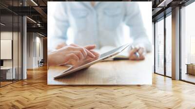 Two female businesswomen working their hands on a tablet in an office in the sunlight at a wooden table and one of them writes a pen in a stylish notebook, and touches the tablet with her finger Wall mural