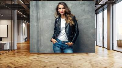 Stylish woman confidently poses in a sleek, black leather jacket, cinched at the waist, over a casual white top and distressed denim jeans. Wall mural