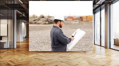 Back view of building engineer standing on construction site, talking by VHF walkie talkie and holding blueprints. Wall mural