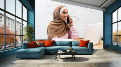 Pretty Muslim woman in hijab is sitting in the classroom at the desk talking on the phone typing on the computer planning and making a school schedule for students. Secretary answering the phone. Wall mural