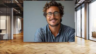 Smiling young Caucasian man with glasses and wavy hair in a denim shirt, against a gray background Wall mural