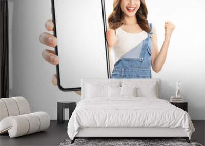 Cheerful young Asian woman holding smartphone with fists clenched celebrating victory expressing success on white background. Wall mural