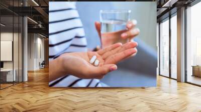 Woman holding white pill on hand and drinking water in glass on sofa in house, feels like sick. Health care concept. Wall mural