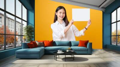 Young Asian woman holding blank paper with smiling face and looking on the yellow background. for advertising signs. Wall mural