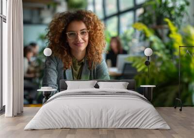 Portrait of a smiling curly-haired businesswoman wearing glasses, with a green office space filled with plants in the background, confident businesswoman smiling in office environment. Wall mural