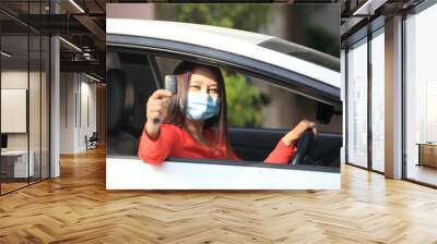  Young woman with medical protective mask on her face is driving a car, she looks into camera and View from outside , Protect yourself. Wall mural