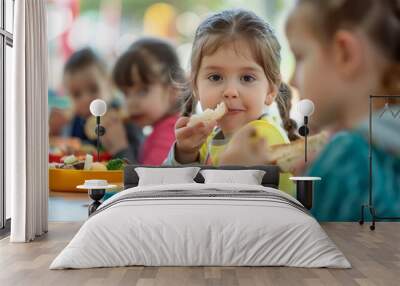 Little girl eating a piece of bread while sitting at a table with other children in the background Wall mural