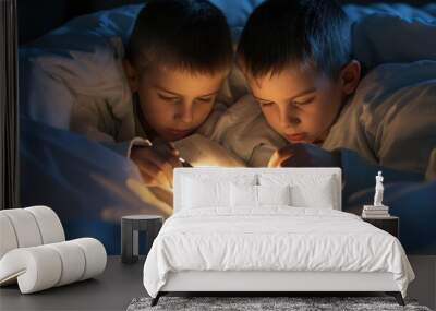 two boys reading a book under a blanket with a flashlight. The light from the flashlight illuminates the page in front of them. They look happy and excited. Halloween Day concept Wall mural