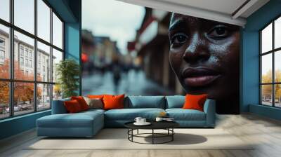 Close-up of a woman with wet skin, looking thoughtful in a rainy city street, with blurred buildings and soft evening light. Wall mural