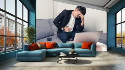 Young Asian business man sitting on sofa and working from home. Man feeling stress and suffering from headache and stomachache. Tired young Asian entrepreneur manager. Work from home concept Wall mural