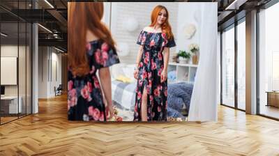 Attractive young woman in floral print dress stands in front large mirror in graceful floral interior. pretty girl with curly red hair, in beautiful dress, in floral interior, smiles. spring festival Wall mural