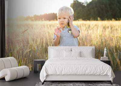 Adorable child talking on a mobile phone, standing in a field of rye in the rays of the setting sun Wall mural