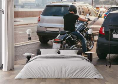 rear view of a handsome biker dressed in a black T-shirt and black helmet jeans and sneakers. He is riding on cruiser motorcycle on background of 2 cars. Traffic stop Wall mural