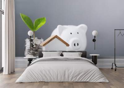 Glass jar filled with coins and a small seedling in the center, with stacked coins white piggy bank and a wooden house model on a table. Saving for house and financial growth Wall mural