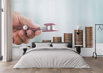 Hand holding a stethoscope, examining a stack of coins on a table against a bright white background. The image symbolizes the concept of a financial health check and monetary well-being. Wall mural