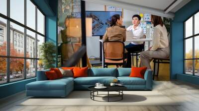 A woman is standing in front of a white board with a smile on her face. She is talking to two other people, one of whom is sitting in a chair. Concept of collaboration and teamwork Wall mural