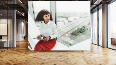 A beautiful African-American woman entrepreneur in a white blouse and red skirt is holding a digital tablet pc while sitting indoors on the bench on the top floor of a business office skyscraper Wall mural