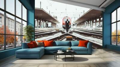 A black stylish bearded bald businessman is walking on the train tracks towards the camera with two symmetrical railroad platforms on the sides and a modern red locomotive in the background Wall mural